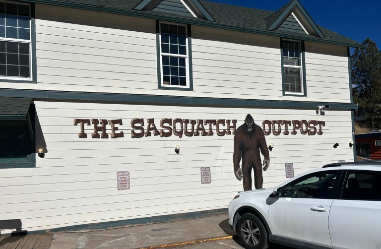The exterior of a building painted white with a sign that says "The Sasquatch Outpost." In between the last two words stands a tall cutout of Sasquatch.