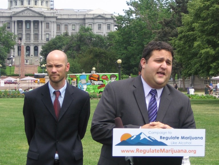 Two men in suits, Brian Vicente and Mason Tvert, stand at a podium in front of the Colorado Capitol Building.