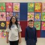 Two young children stand in front of a wall decorated with children's artwork.