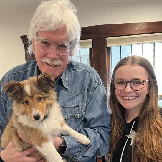 An older man with white hair and a white mustache holds a puppy and stands next to a young woman.