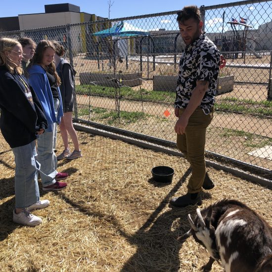 A man stands in front of several students pointing at a goat.